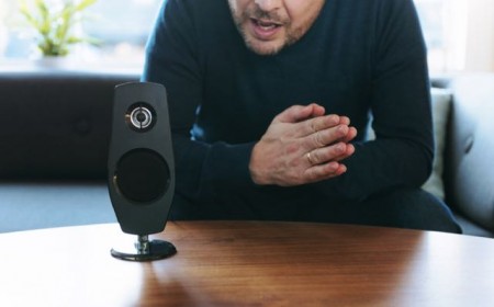 Man sits in his living room and asks a question to his digital assistant placed on a table. The device is connected to the internet and has both a microphone and a speaker. It recieves the questions and provide answers. It can also carry out different tasks as turning on the lights and music at home by voice command.