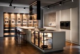 Im Mittelpunkt des Interieurs steht die großvolumige Insel, deren monolithischer Charakter aufgelöst wird durch die Transparenz der beleuchteten Unterschrank-Vitrinen mit schwarzmatt gebürsteten Metallrahmen. Die Strenge der Formensprache und Materialität durchbricht ein warmer Akzent: die auf die filigrane Ceramic-Arbeitsplatte aufgesetzte Bartheke mit ihrer elliptischen Rundung und ihrer Oberfläche aus hellem Holz. © Copyright SieMatic