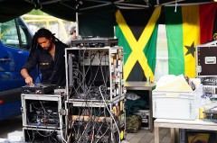 Ein DJ bedient unter einem Pavillon ein Soundsystem mit Plattenspieler und umfangreicher Tontechnik; im Hintergrund hängen die Flaggen Jamaikas und Ghanas.