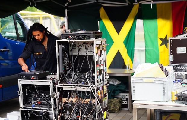 Ein DJ bedient unter einem Pavillon ein Soundsystem mit Plattenspieler und umfangreicher Tontechnik; im Hintergrund hängen die Flaggen Jamaikas und Ghanas.