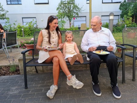 Bewohner, Angehörige und Kinder sitzen gemeinsam im Garten und essen bei einem Sommerfest