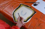 Hand mit Pinsel beim Bemalen eines mittelalterlichen Schildmotivs auf einem orangefarbenen Basteltisch.