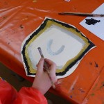 Hand mit Pinsel beim Bemalen eines mittelalterlichen Schildmotivs auf einem orangefarbenen Basteltisch.