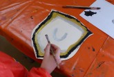 Hand mit Pinsel beim Bemalen eines mittelalterlichen Schildmotivs auf einem orangefarbenen Basteltisch.
