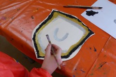 Hand mit Pinsel beim Bemalen eines mittelalterlichen Schildmotivs auf einem orangefarbenen Basteltisch.