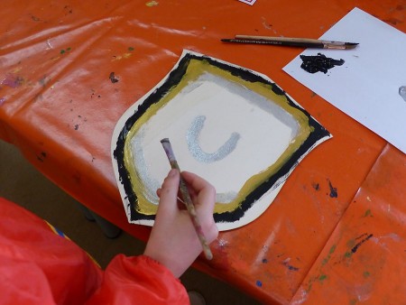 Hand mit Pinsel beim Bemalen eines mittelalterlichen Schildmotivs auf einem orangefarbenen Basteltisch.