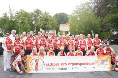 Gruppe von Radfahrerinnen und Radfahrern in roten Trikots der Radtour pro Organspende mit Banner bei einem Halt in Gütersloh
