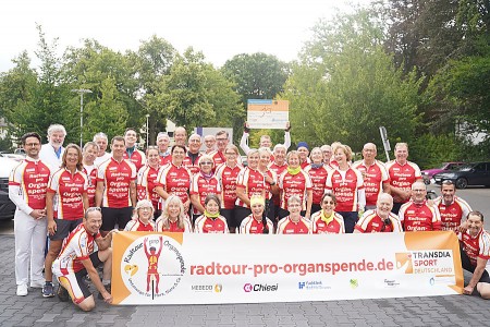 Gruppe von Radfahrerinnen und Radfahrern in roten Trikots der Radtour pro Organspende mit Banner bei einem Halt in Gütersloh