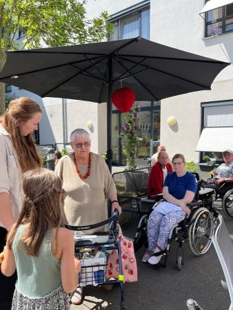 Seniorinnen und Senioren sowie Kinder treffen sich im Garten eines Pflegeheims unter einem Sonnenschirm