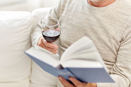 Ein älterer Mann sitzt auf einem Sofa, liest ein Buch und hält ein Glas Rotwein in der Hand.