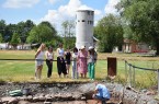 Besuchergruppe beobachtet eine archäologische Grabung auf einer Grünfläche; im Hintergrund steht ein weißer Turm.
