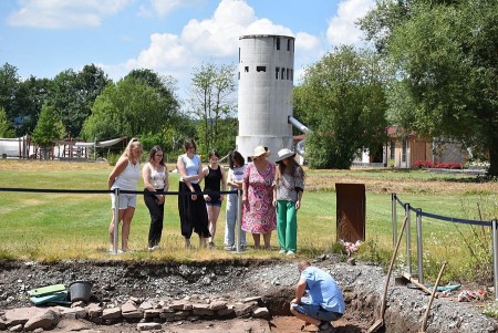 Besuchergruppe beobachtet eine archäologische Grabung auf einer Grünfläche; im Hintergrund steht ein weißer Turm.