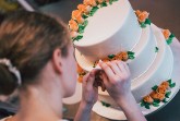 Eine Person dekoriert eine dreistöckige weiße Torte mit orangefarbenen Rosen und grünen Blättern.