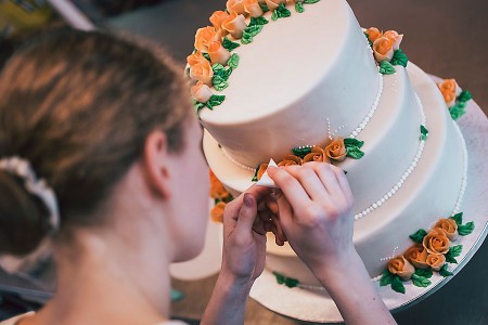 Eine Person dekoriert eine dreistöckige weiße Torte mit orangefarbenen Rosen und grünen Blättern.