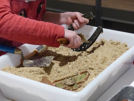 Kinderhände graben mit kleinen Werkzeugen in einer mit Sand gefüllten Ausgrabungsstation Vorhandener © Mindener Museum