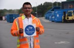 Mann in orangefarbener Warnjacke hält auf einem Recyclinghof ein Schild mit der Aufschrift „Bargeldlose Zahlung bevorzugt!“.