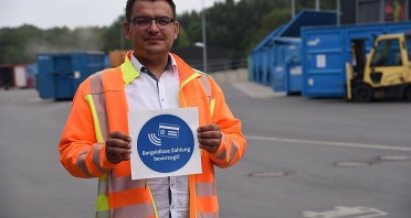 Mann in orangefarbener Warnjacke hält auf einem Recyclinghof ein Schild mit der Aufschrift „Bargeldlose Zahlung bevorzugt!“.