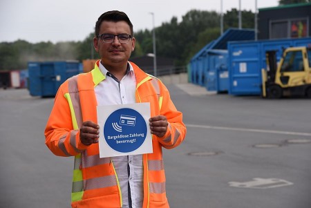 Mann in orangefarbener Warnjacke hält auf einem Recyclinghof ein Schild mit der Aufschrift „Bargeldlose Zahlung bevorzugt!“.