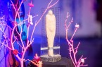 Figurförmige Skulptur der BigBrotherAwards vor einer transparenten Zahlentafel, umgeben von pink beleuchteten Zweigen..