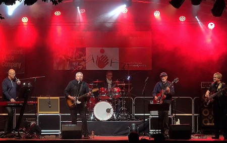 Fünfköpfige Rockband mit Gitarren, Keyboard und Schlagzeug bei einem Bühnenauftritt unter rotem Licht.