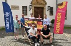 Sieben Beteiligte präsentieren das Plakat zum Open-Air-Sommer in Büren zwischen zwei Veranstaltungsfahnen vor dem Rathaus.