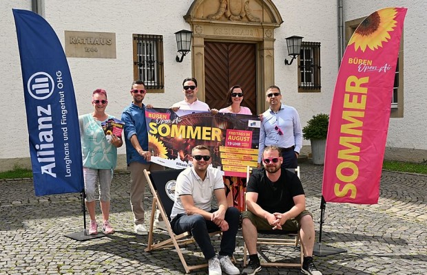 Sieben Beteiligte präsentieren das Plakat zum Open-Air-Sommer in Büren zwischen zwei Veranstaltungsfahnen vor dem Rathaus.