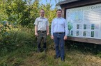 Zwei Männer stehen im Bürgerwald Extertal neben einer Informationstafel, umgeben von dichtem Bewuchs.