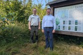 Zwei Männer stehen im Bürgerwald Extertal neben einer Informationstafel, umgeben von dichtem Bewuchs.