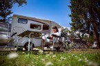 Frau mit Laptop in einem Campingstuhl vor einem Wohnmobil; daneben stehen zwei Fahrräder auf einem bewaldeten Campingplatz.