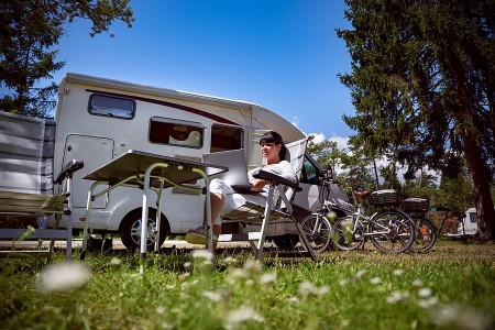 Frau mit Laptop in einem Campingstuhl vor einem Wohnmobil; daneben stehen zwei Fahrräder auf einem bewaldeten Campingplatz.