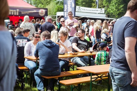 Besucher sitzen und stehen tagsüber zwischen Biertischen und Foodtrucks auf einem Streetfoodfestival.
