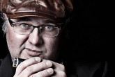 Nahaufnahme eines Mannes mit brauner Schirmmütze und Brille, der eine Mundharmonika in der Hand hält, vor schwarzem Hintergrund.