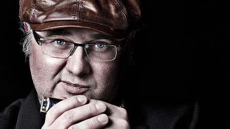 Nahaufnahme eines Mannes mit brauner Schirmmütze und Brille, der eine Mundharmonika in der Hand hält, vor schwarzem Hintergrund.