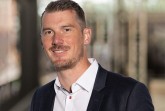 Christopher Boss im dunklen Sakko und weißen Hemd vor einem unscharfen Messehintergrund.