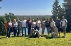 14 Mitglieder der Delegationen stehen und sitzen auf einer Wiese vor der bewaldeten Seenlandschaft am Aussichtshügel Ladakalnis.