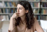 Junge Frau sitzt mit einem Stift in der Hand vor einem Bücherregal und blickt nachdenklich zur Seite.
