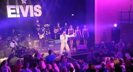 Elvis-Performer in weißem Bühnenanzug bei einem Konzert mit Band und Backgroundsängerinnen vor Publikum.
