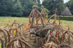 Mehrere aus Stroh gebundene Erntekronen mit bunten Bändern auf einer Wiese; im Hintergrund stehen Bäume und ein Gebäude.