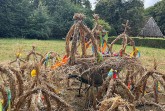 Mehrere aus Stroh gebundene Erntekronen mit bunten Bändern auf einer Wiese; im Hintergrund stehen Bäume und ein Gebäude.