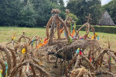Mehrere aus Stroh gebundene Erntekronen mit bunten Bändern auf einer Wiese; im Hintergrund stehen Bäume und ein Gebäude.