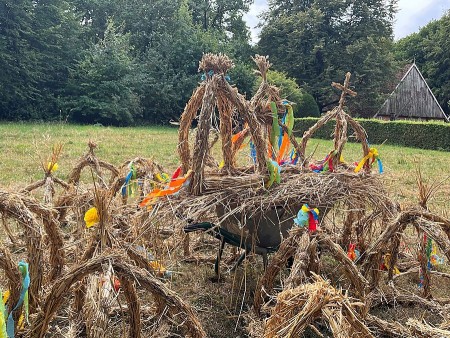 Mehrere aus Stroh gebundene Erntekronen mit bunten Bändern auf einer Wiese; im Hintergrund stehen Bäume und ein Gebäude.
