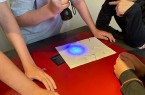 Mehrere Kinder und Jugendliche stehen um einen roten Tisch und untersuchen Papier-Rätsel mit einer UV-Taschenlampe im Mindener Museum.