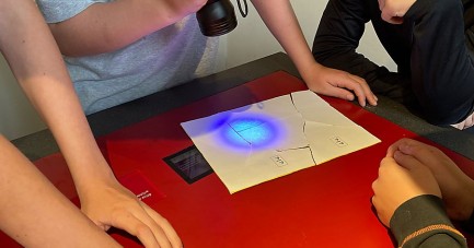Mehrere Kinder und Jugendliche stehen um einen roten Tisch und untersuchen Papier-Rätsel mit einer UV-Taschenlampe im Mindener Museum.
