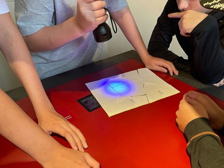 Mehrere Kinder und Jugendliche stehen um einen roten Tisch und untersuchen Papier-Rätsel mit einer UV-Taschenlampe im Mindener Museum.