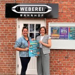 Doreen Belhaouès und Jana Felmet stehen vor dem Weberei-Bahnhof und halten ein Plakat zum Kinderprogramm.