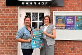 Doreen Belhaouès und Jana Felmet stehen vor dem Weberei-Bahnhof und halten ein Plakat zum Kinderprogramm.