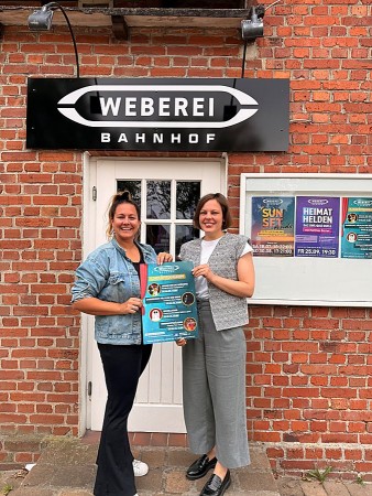 Doreen Belhaouès und Jana Felmet stehen vor dem Weberei-Bahnhof und halten ein Plakat zum Kinderprogramm.
