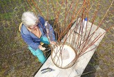 Eine Frau flechtet im Freien an einem Holztisch einen großen Korb aus langen Weidenzweigen.