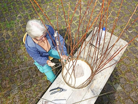 Eine Frau flechtet im Freien an einem Holztisch einen großen Korb aus langen Weidenzweigen.