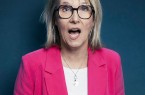 Blonde Frau mit schwarzer Brille und pinkfarbenem Blazer vor dunkelblauem Hintergrund.